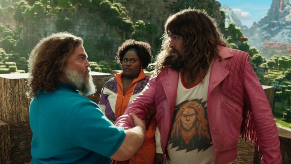 Jack Black shakes hands with Jason Momoa, who wears a bright pink jacket, while Danielle Brooks looks on in a vivid outdoor Minecraft landscape. - Cast of Minecraft The Movie