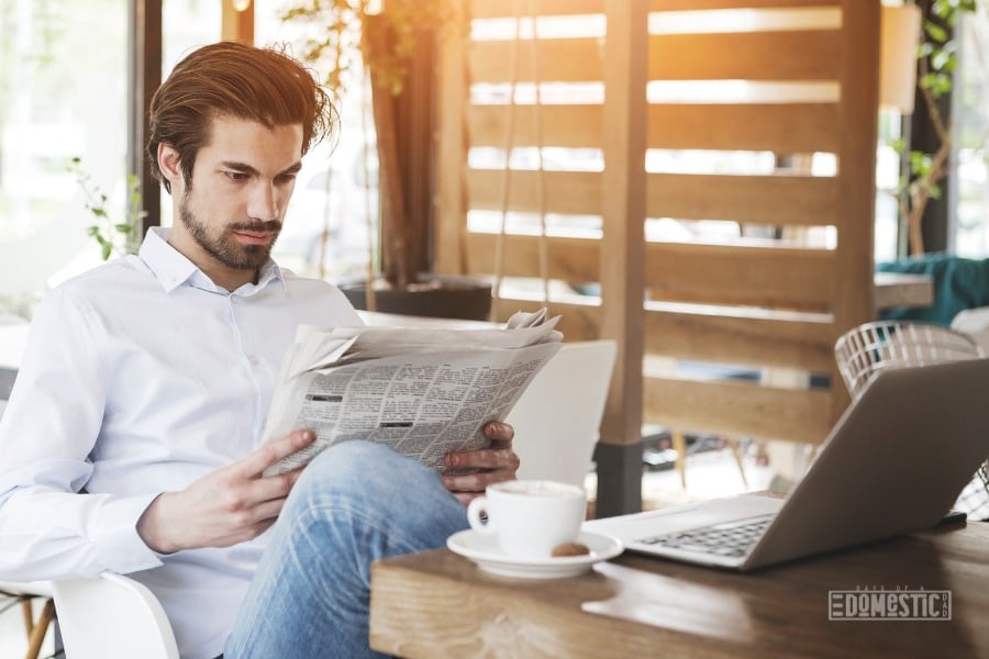 man reading a newspaper with cup of coffee and laptop on table in front of him