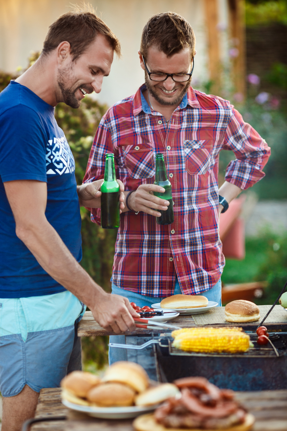 Two men enjoying a backyard barbecue, both holding beers and smiling as they grill food. One man is wearing a blue t-shirt and light-colored shorts, while the other sports a classic BBQ dad outfit with a red plaid shirt and jeans. They stand near a grill with corn and other food items, embodying a relaxed and cheerful summer vibe.