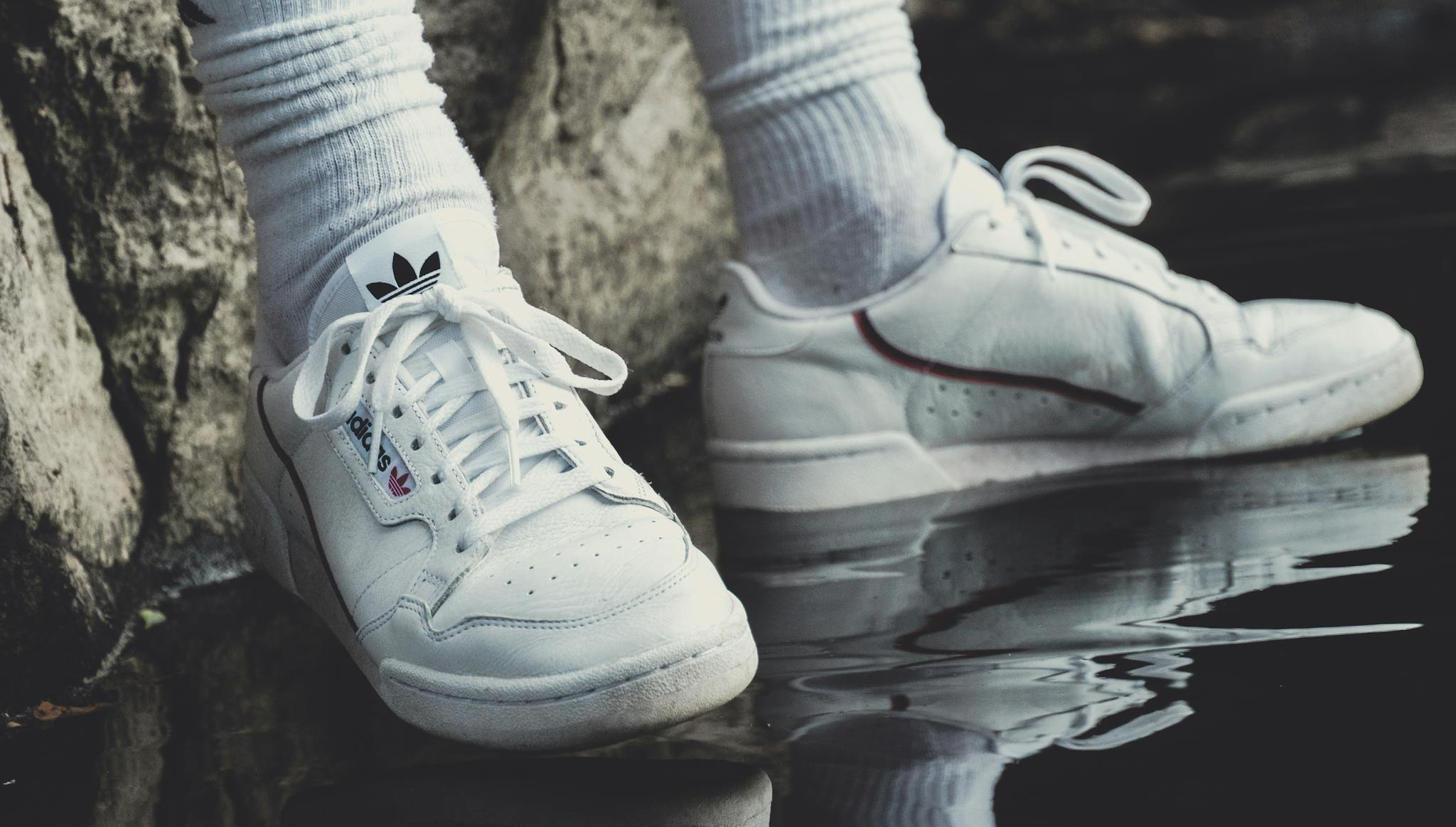 Close-up of white sneakers and socks reflecting on water next to a rock.