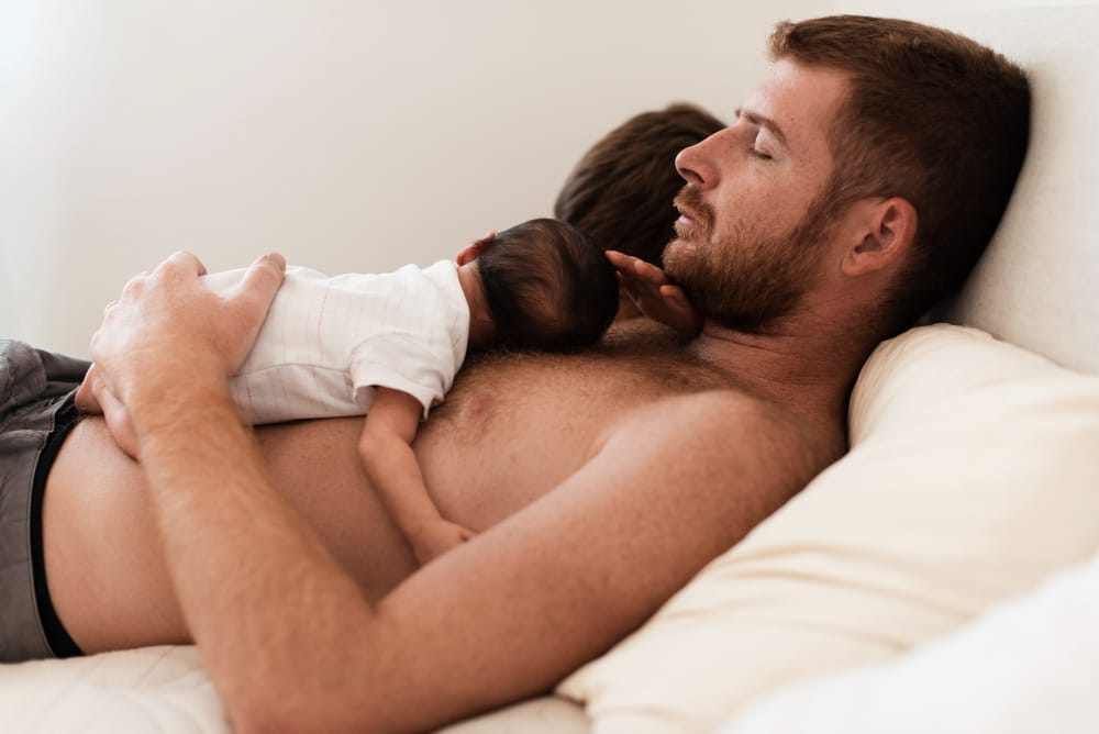 Father lying down with newborn baby sleeping peacefully on his bare chest, depicting intimate bonding moment