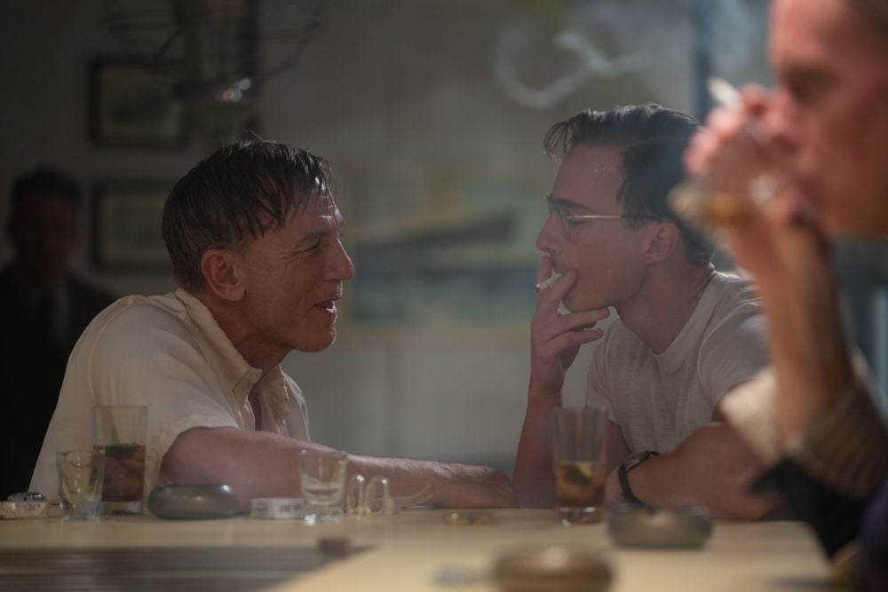 Daniel Craig and Drew Starkey share a conversation and cigarettes at a dimly lit bar.