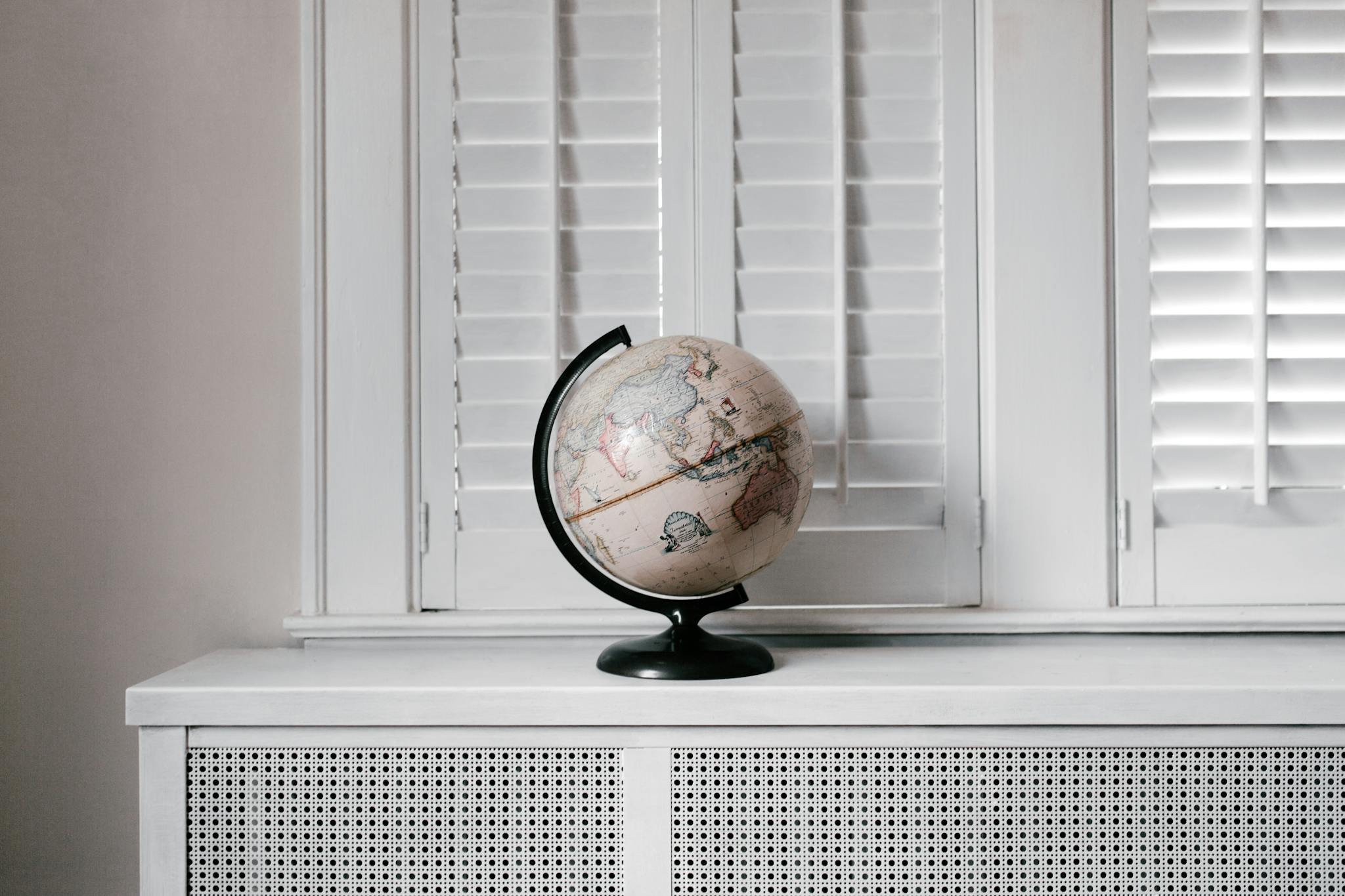 Elegant globe on a minimalist white desk against shuttered windows in modern interior design.