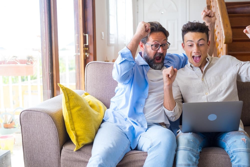 Happy excited grown son and mature 50s father using laptop, celebrating success. two family generations of sport fan watching online game or match on computer, making yes win gesture, having fun