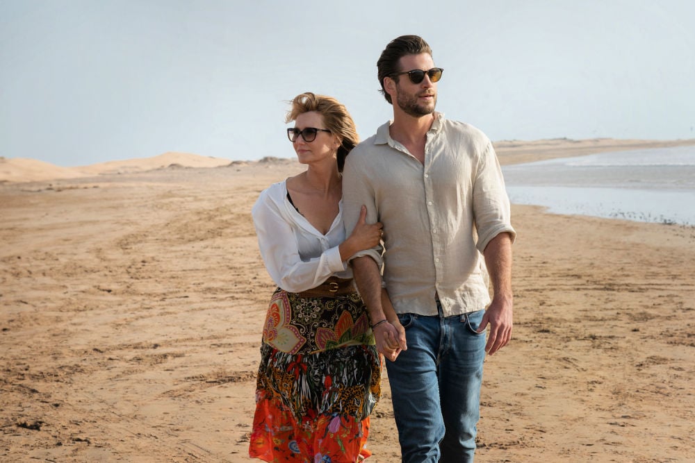 Laura Dern and Liam Hemsworth walking hand in hand along a serene beach, portraying their characters Katherine and Owen in a scene from the Netflix romantic drama Lonely Planet