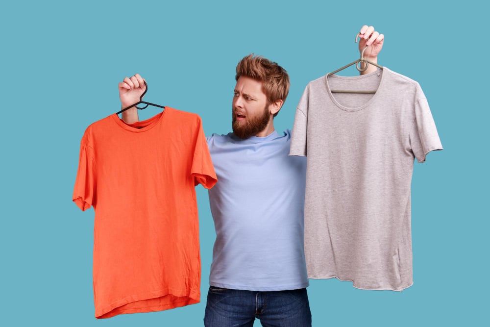 Man frowning face holding two hangers with gray and orange tshirts hard to choose the best attire