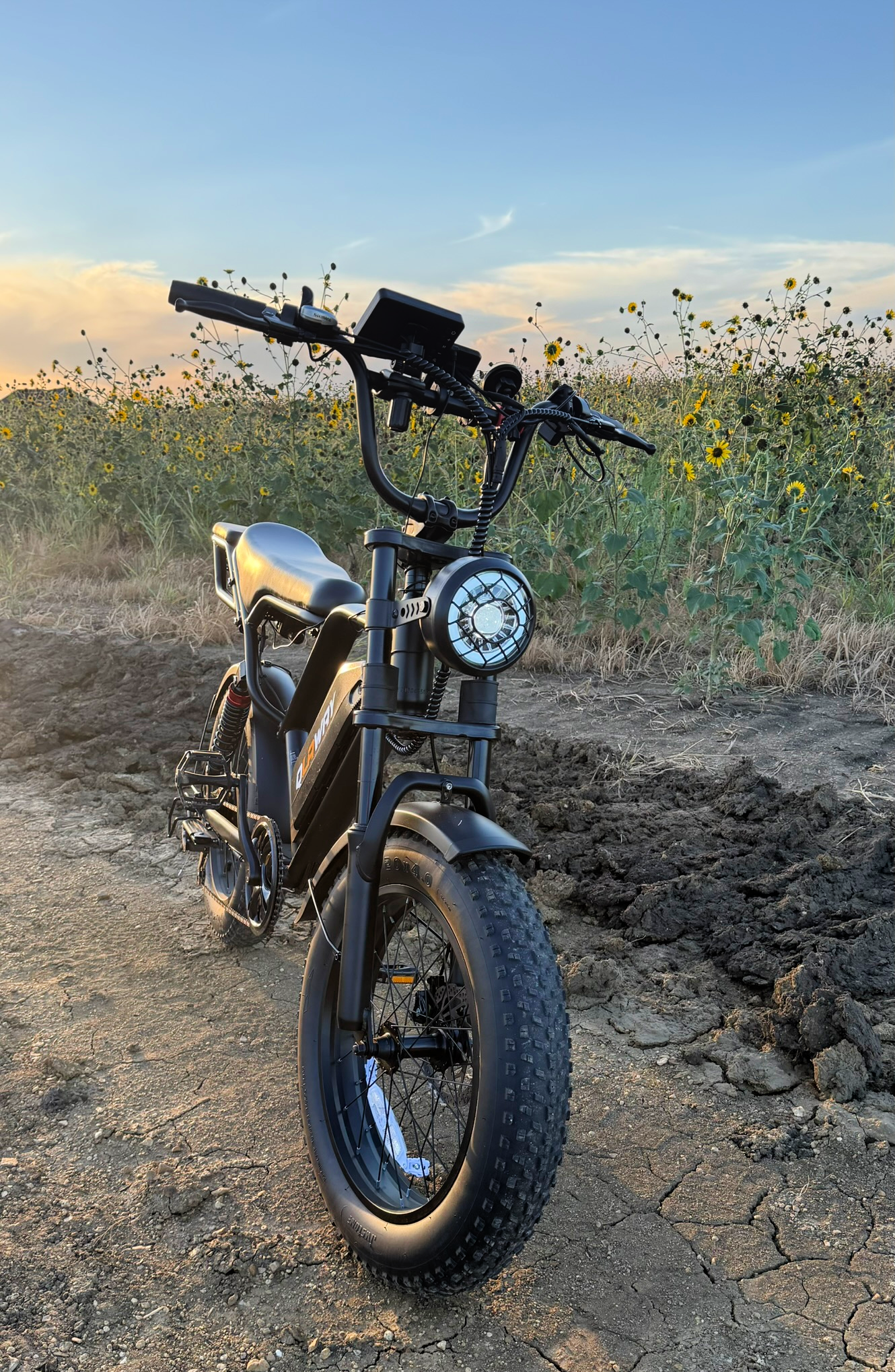 Front three-quarter view of a moped-style fat-tire e-bike with round headlight at sunset.