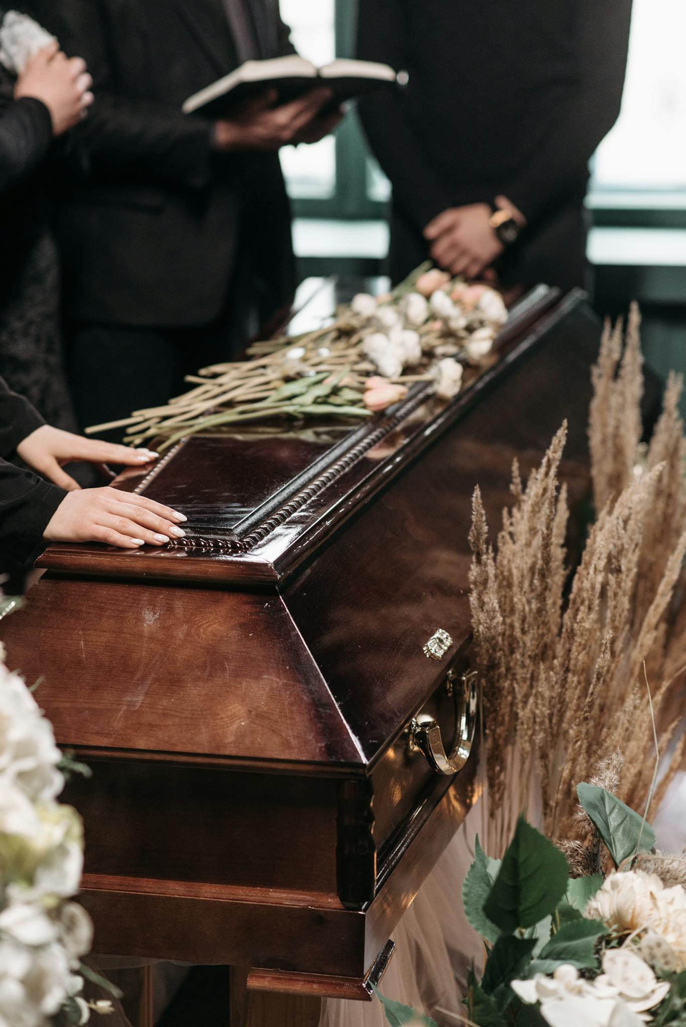 Mourners at a funeral with floral arrangements on a polished wooden coffin.