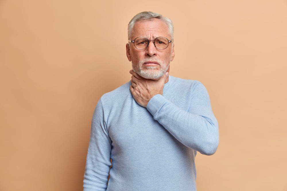 Free photo old handsome bearded man touches neck suffocates because of painful strangle feels pain in throat while swallow wears casual jumper isolated over beige studio wall

