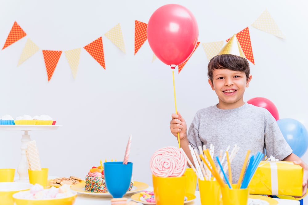 Free photo smiling boy wearing party hat holding balloon and gift standing behind variety of food on table