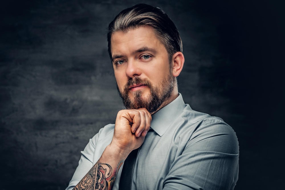 stylish bearded male with tattooed arms, dressed in a shirt posing over grey background