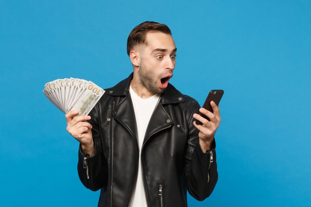Young man in black leather jacket white t-shirt holding fan of cash money in dollar banknotes, cellphone isolated on blue wall background studio portrait. people lifestyle concept. mock up copy space.