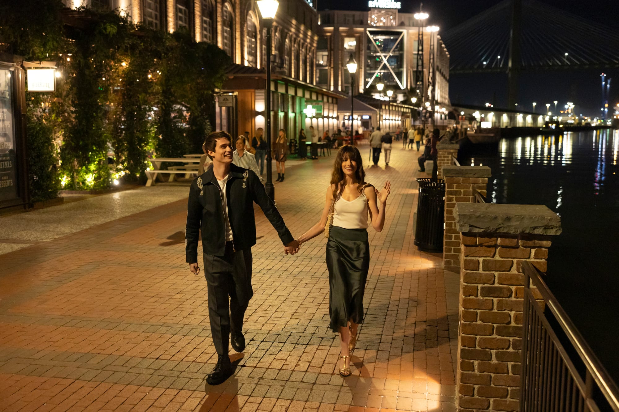 A couple holding hands while walking along a waterfront promenade at night in a scene from The Idea of You.