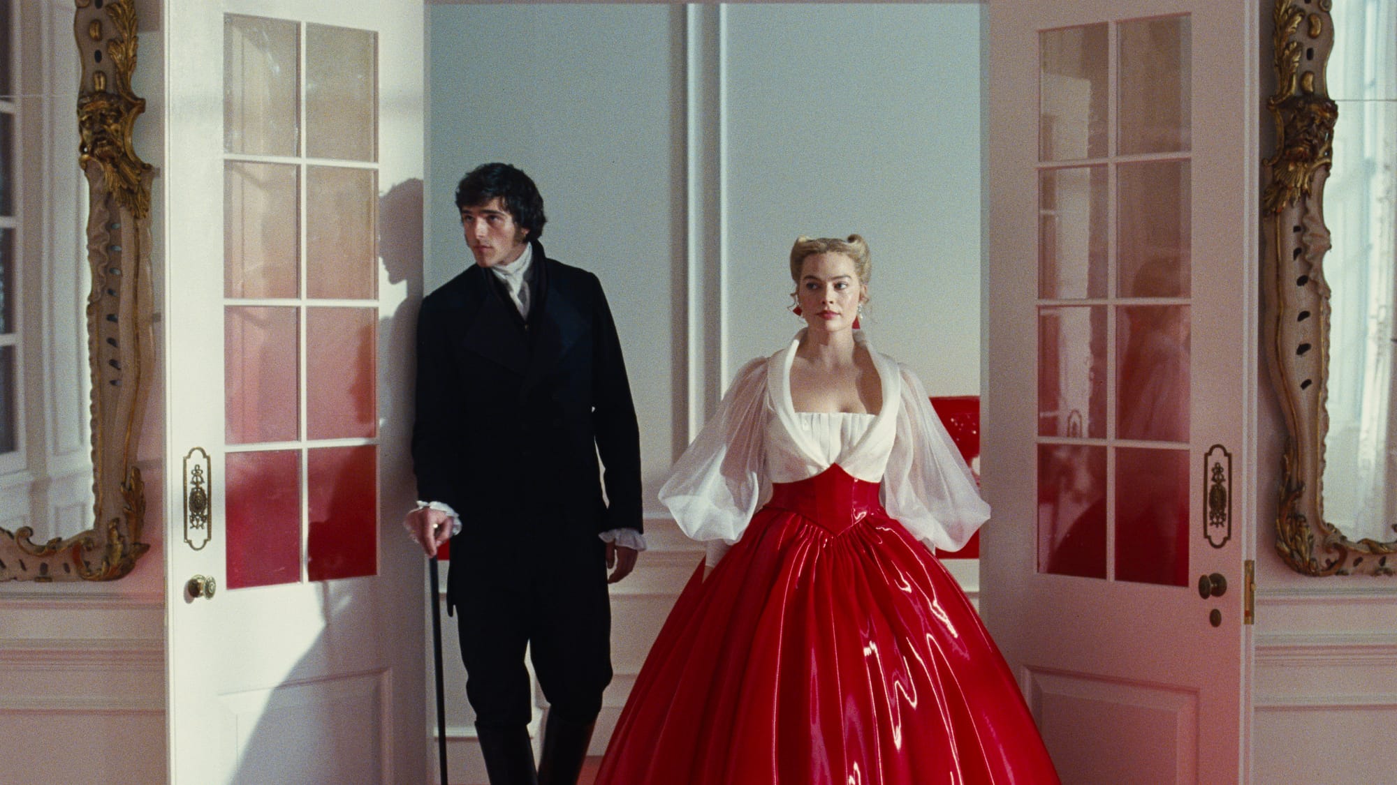 Margot Robbie in a dramatic red dress beside Jacob Elordi in a doorway scene from Wuthering Heights (2026), illustrating how the film’s beauty can mask unhealthy and obsessive relationships.