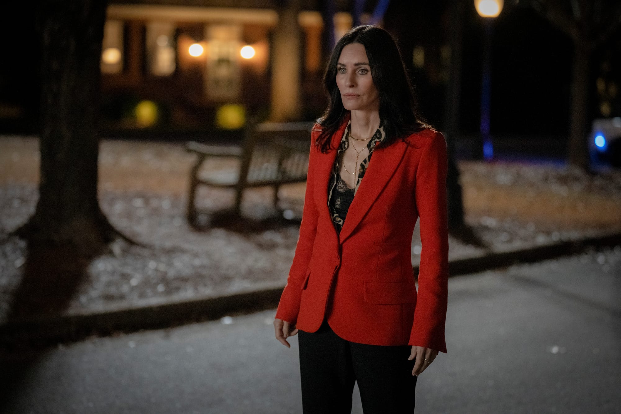 Gale Weathers (Courteney Cox) on a dark street at night, standing and looking contemplative. She is wearing a bold red blazer and black pants.