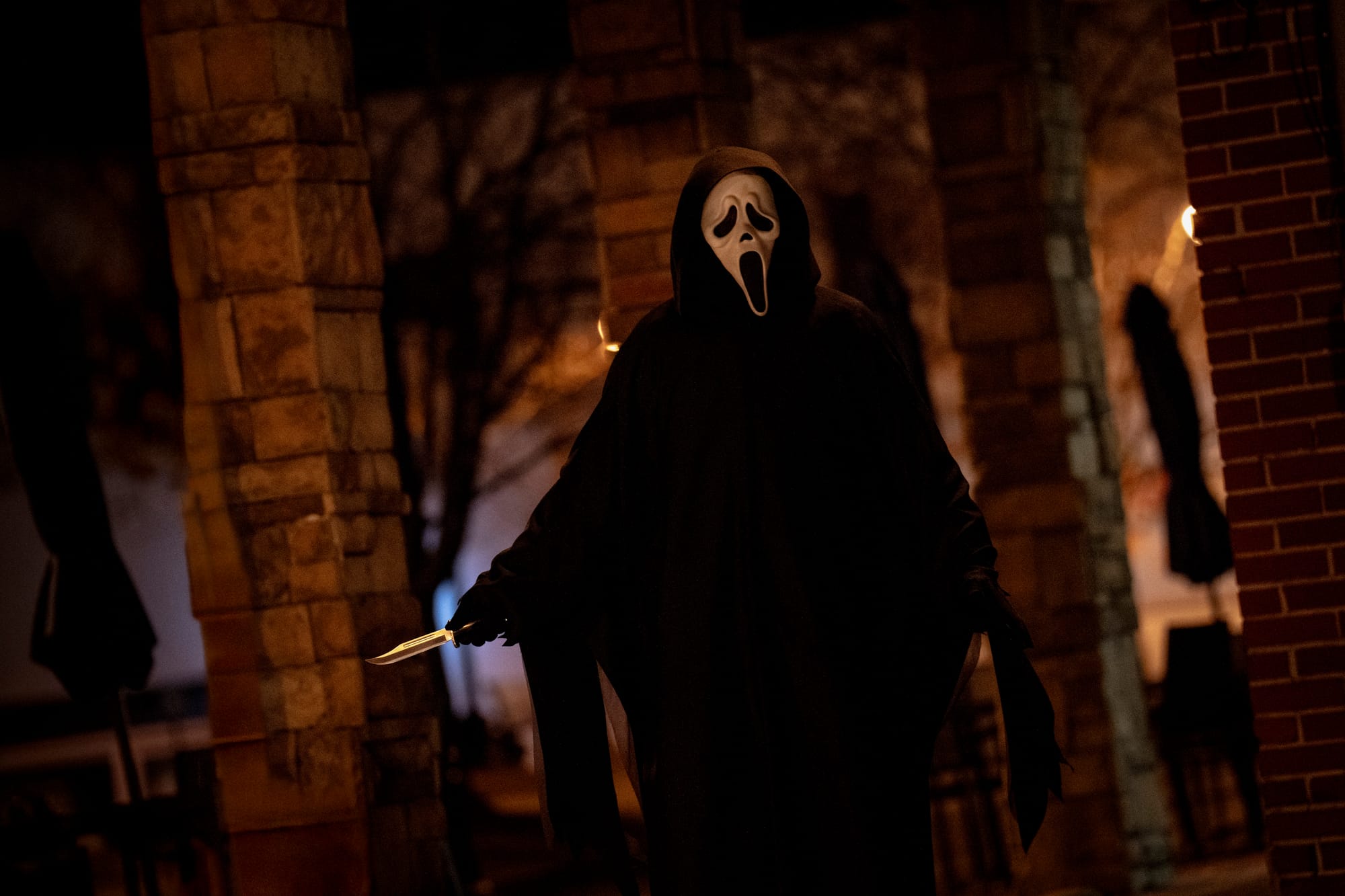 Full-body movie still with Ghostface standing in a dark alleyway at night, wearing a black robe and mask, and holding a large hunting knife.