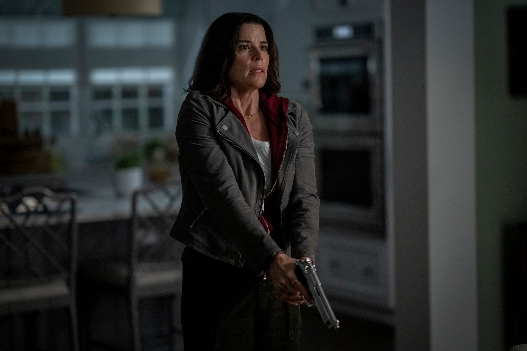 Sidney Prescott (Neve Campbell) in a dimly lit kitchen, holding a large silver semi-automatic handgun and looking to the right with an anxious expression.
