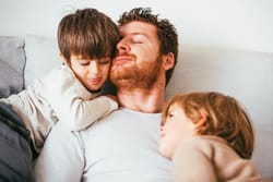 Father relaxing on couch with eyes closed while two young children cuddle close to him, showing peaceful moment of connection and love
