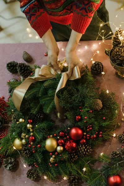 A close-up of hands arranging a diy christmas decorations wreath with ornaments and ribbon on a decorated table.