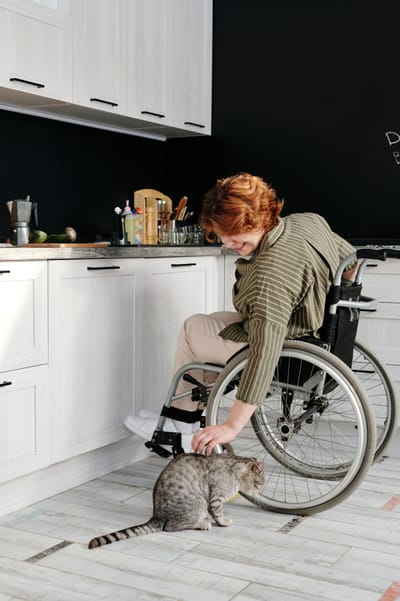 A woman in a wheelchair smiles while petting a tabby cat in a modern kitchen.