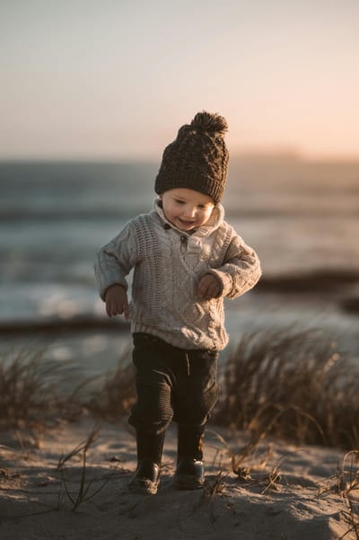 Adorable toddler in warm knitwear exploring a sandy beach with a joyful smile at sunset.