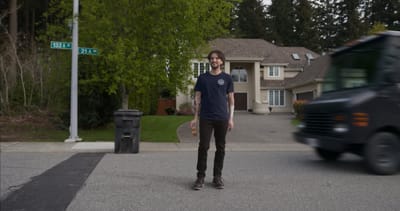 A man smiles while standing casually in a suburban street, unaware of an oncoming delivery truck speeding toward him.