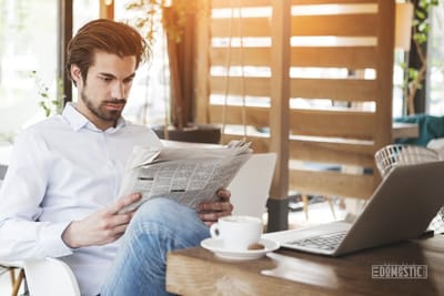 man reading a newspaper with cup of coffee and laptop on table in front of him