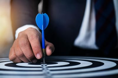 Close-up of a man's hand holding a blue dart on a bullseye, symbolizing precision and focus.