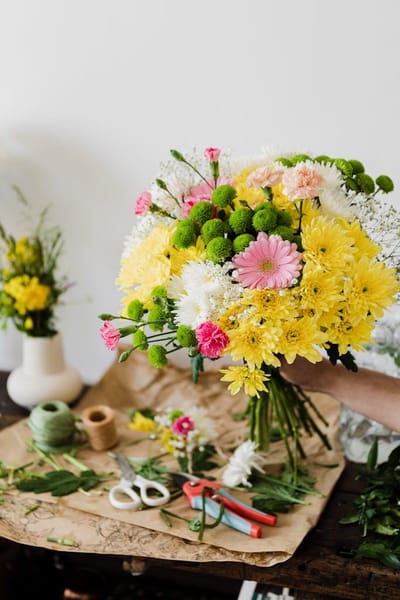 Crop unrecognizable florist composing chrysanthemum bouquet in workshop near messy table covered with cut leaves and floristr