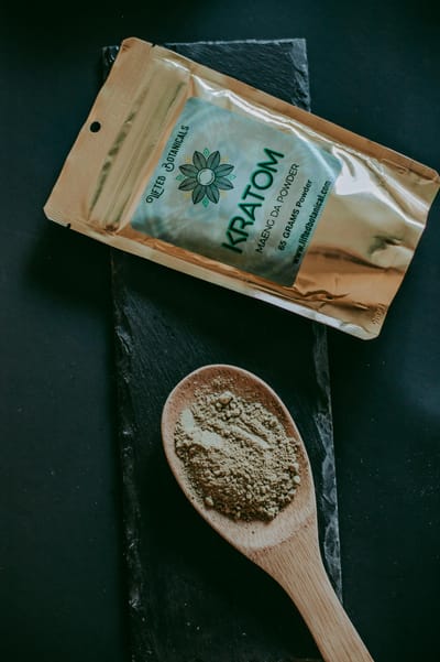 Kratom Maeng Da powder displayed with packet and wooden spoon on a dark surface.