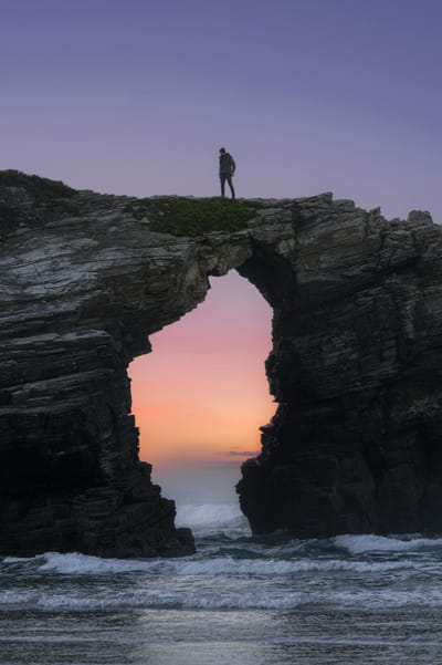 Man stands atop a stone arch overlooking the ocean at sunset in Ribadeo, Spain.