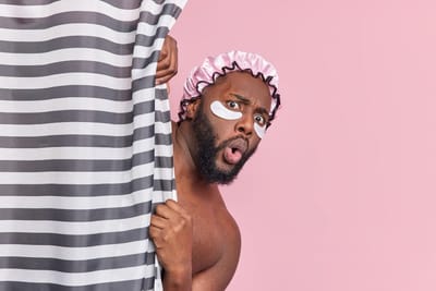 Free photo shocked bearded man hides behind shower curtain has amazed face expression applies beauty patches under eyes has d