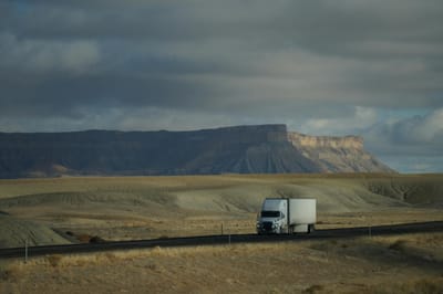 Truck driving through vast desert landscape with dramatic cloudy sky and mountain backdrop.