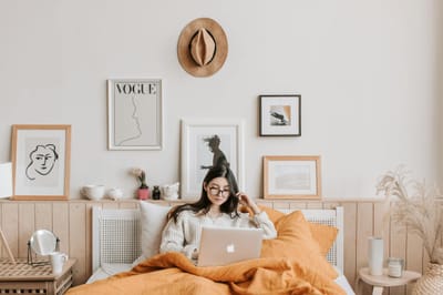 Young woman in cozy bedroom using a laptop, surrounded by stylish decor and art.