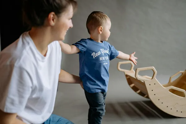 A young boy in a blue shirt playing with a wooden rocking toy under adult supervision indoors.