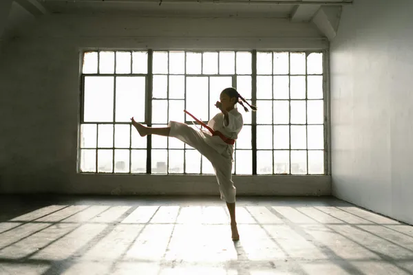 A young girl in a martial arts uniform practicing a high kick in a sunlit dojo.
