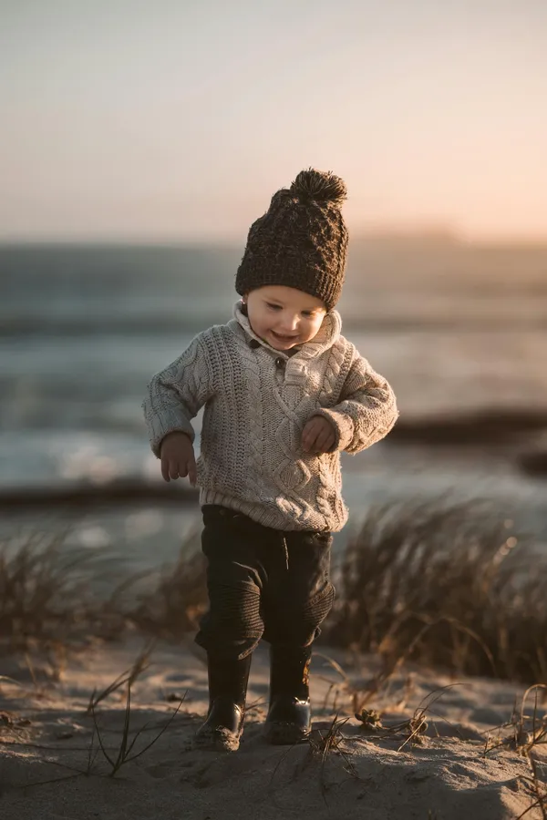 Adorable toddler in warm knitwear exploring a sandy beach with a joyful smile at sunset.
