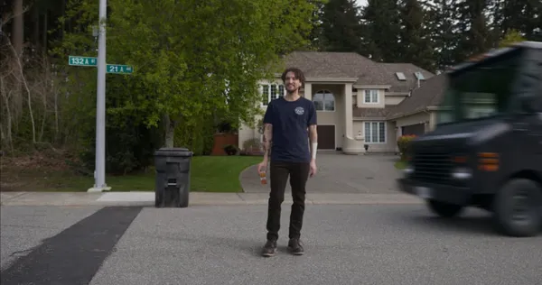 A man smiles while standing casually in a suburban street, unaware of an oncoming delivery truck speeding toward him.