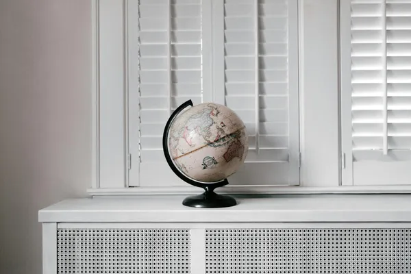 Elegant globe on a minimalist white desk against shuttered windows in modern interior design.
