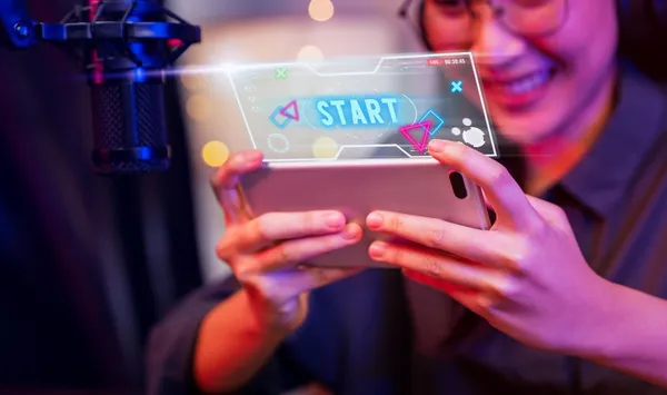 Excited young woman wearing headset and playing online game on smartphone with live broadcasting on internet