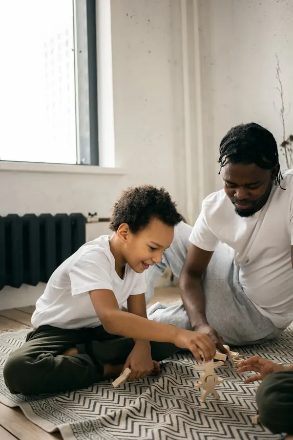 Pensive African American father in casual clothes sitting on floor and playing in board game with little son at home in dayti