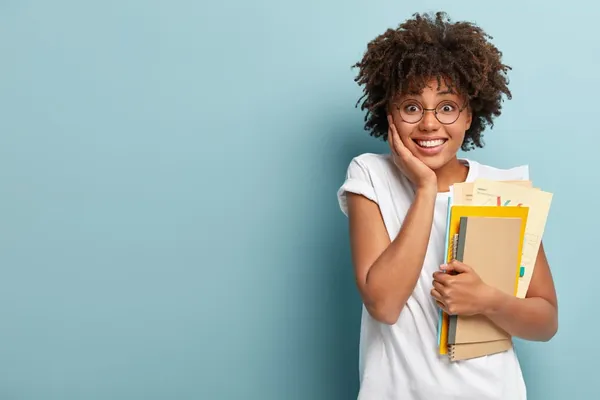 Pleasant looking afro american woman holds notepads, papers, studies at college, glad to finish studying