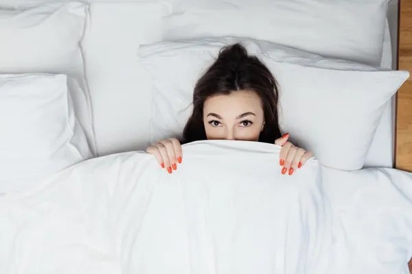 Portrait of surprised lady laying in the bed under white duvet and covers her body