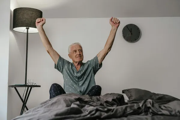 Senior man in pajamas waking up with arms stretched in a cozy bedroom setting.