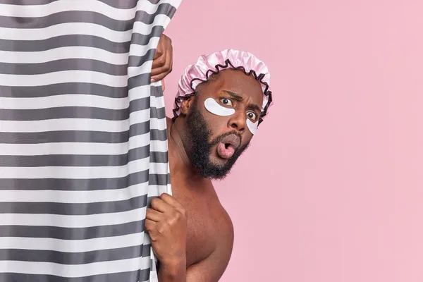 Free photo shocked bearded man hides behind shower curtain has amazed face expression applies beauty patches under eyes has d
