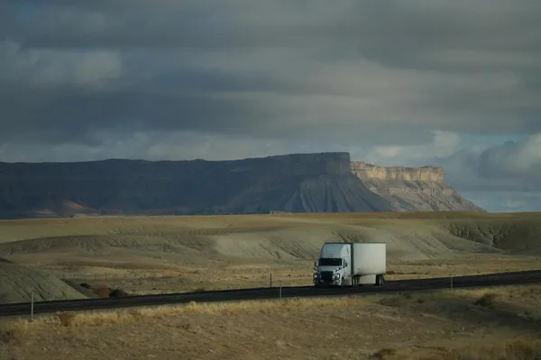 Truck driving through vast desert landscape with dramatic cloudy sky and mountain backdrop.