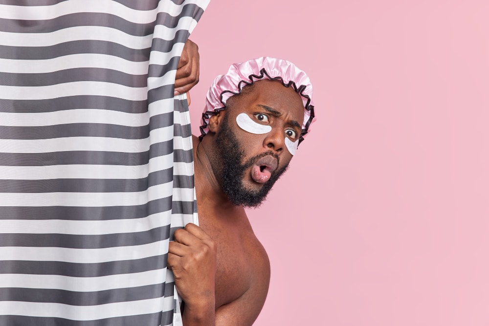 Free photo shocked bearded man hides behind shower curtain has amazed face expression applies beauty patches under eyes has daily hygiene procedures isolated over pink wall