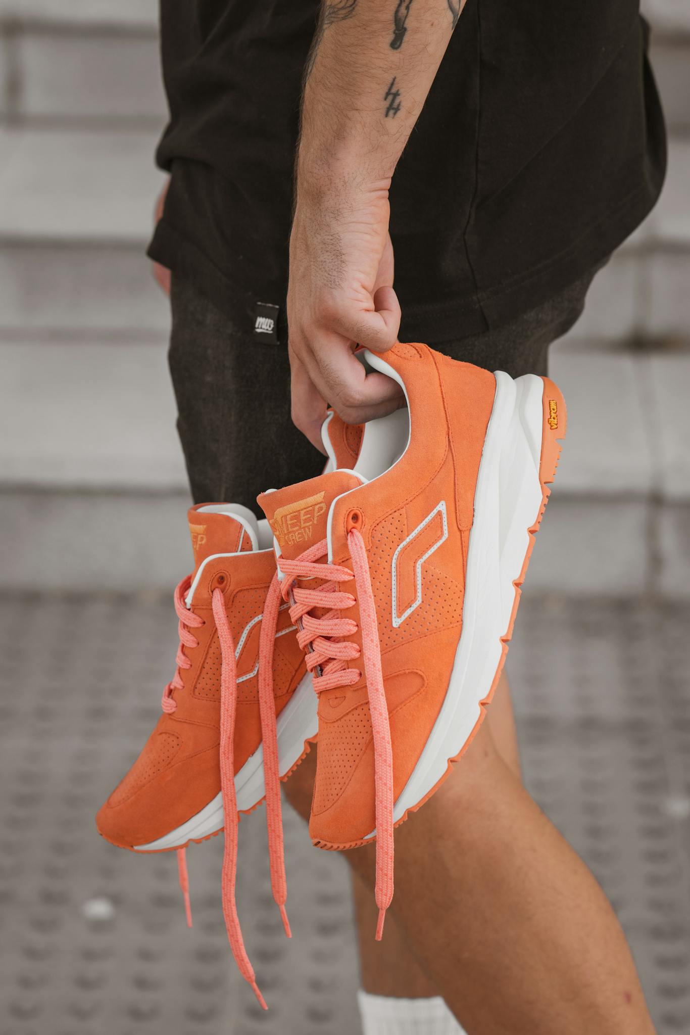 Person holding orange sneakers with tattooed arm, featuring black shorts in close-up.