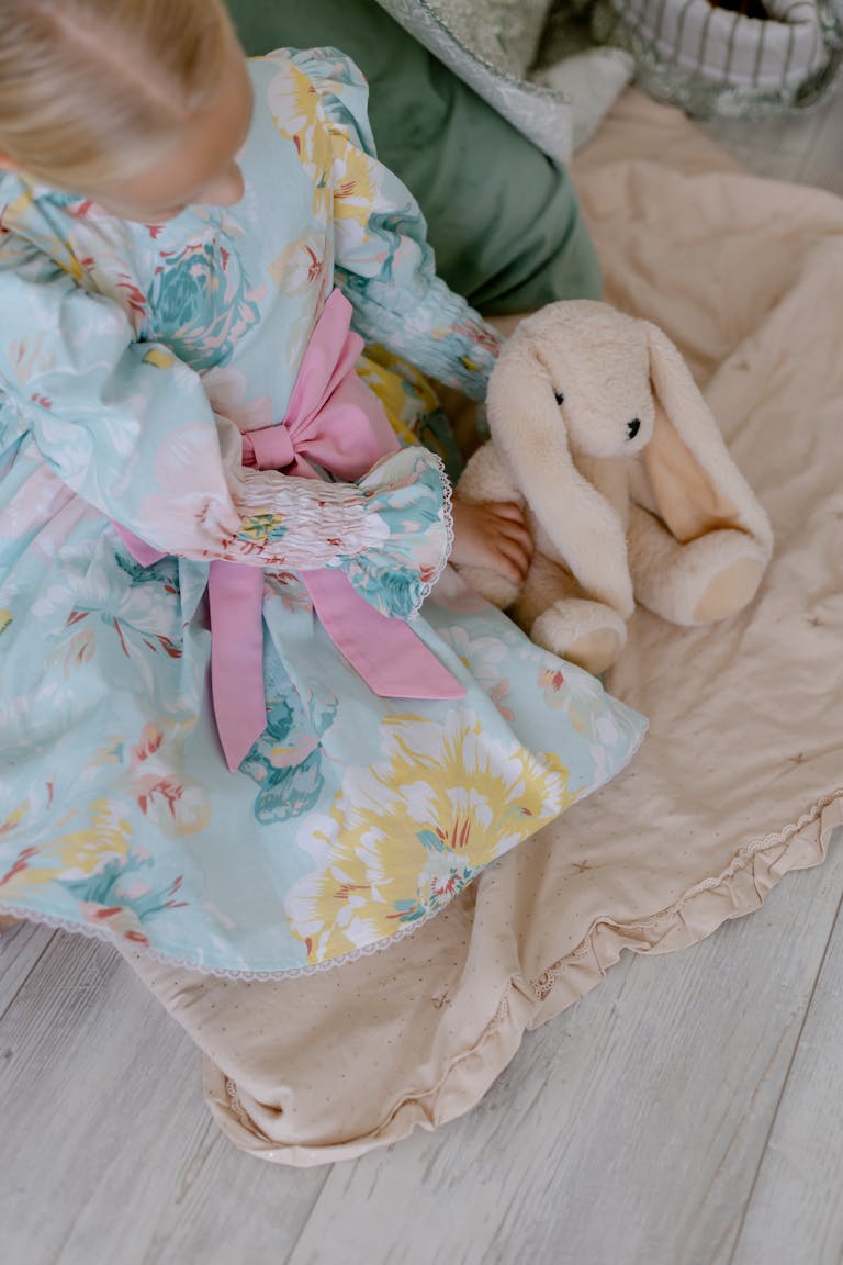 A young girl in a floral dress with a pink bow sits with a plush bunny toy on a cozy beige blanket.