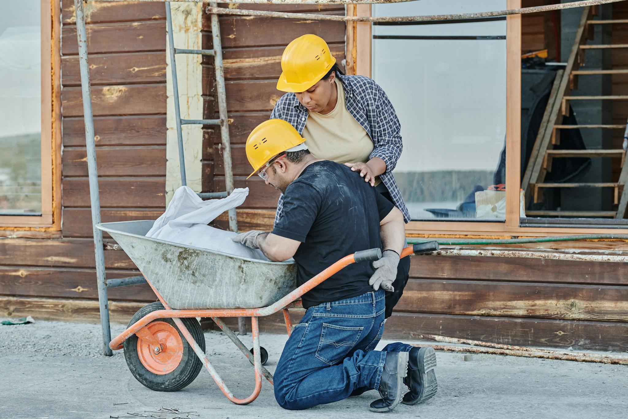 A construction worker receiving help while kneeling with a wheelbarrow on a job site.